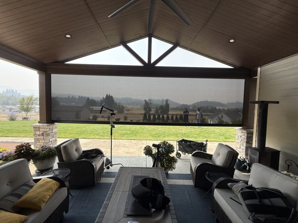 View through a lowered motorized shade screen from inside a covered patio, looking out over farmland and hills